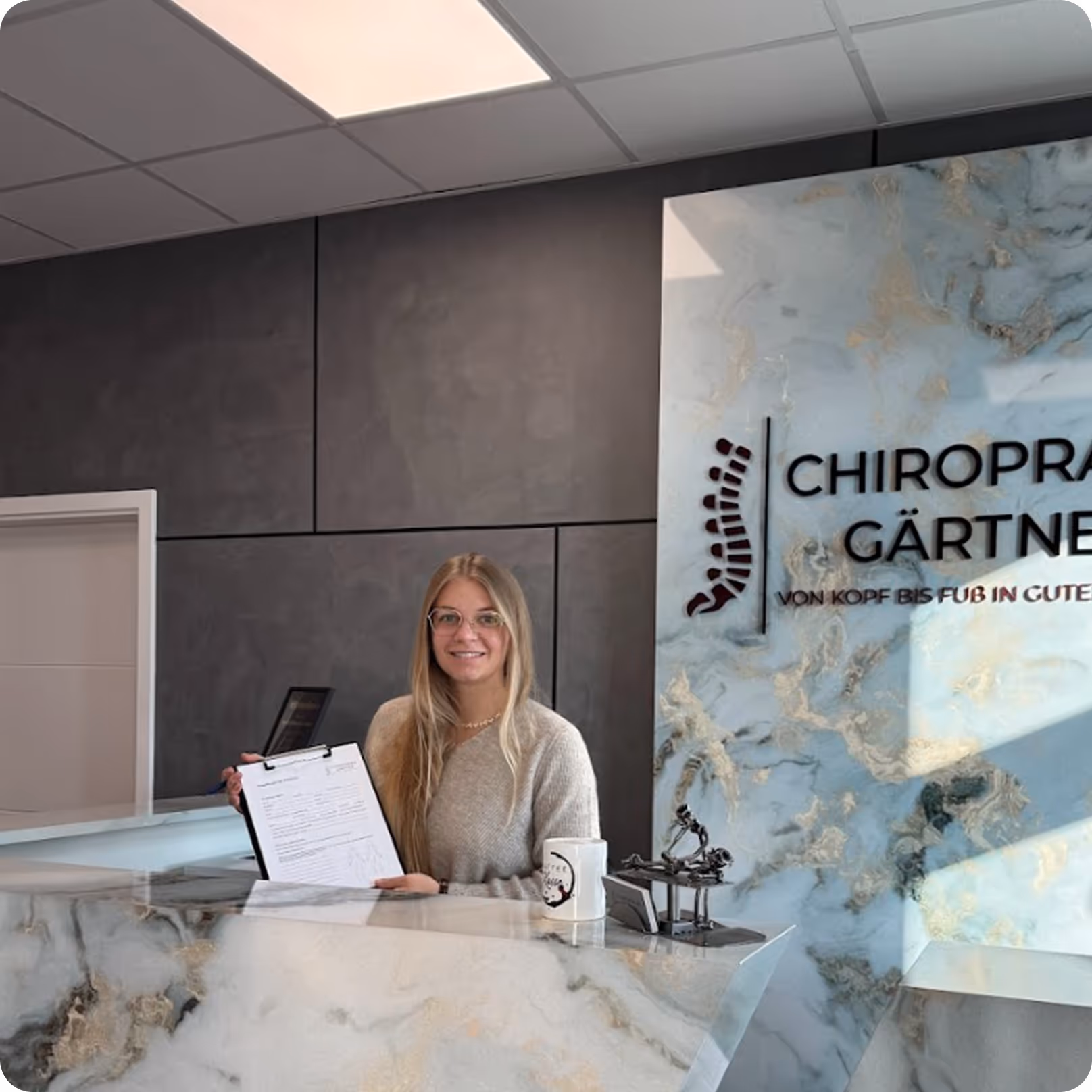 Eine Frau mit langen blonden Haaren und Brille steht hinter einem Marmor-Empfangstresen in einer modernen Praxis von Chiropraxis Sven Gärtner und hält ein Formular auf einer Klemmbrett.