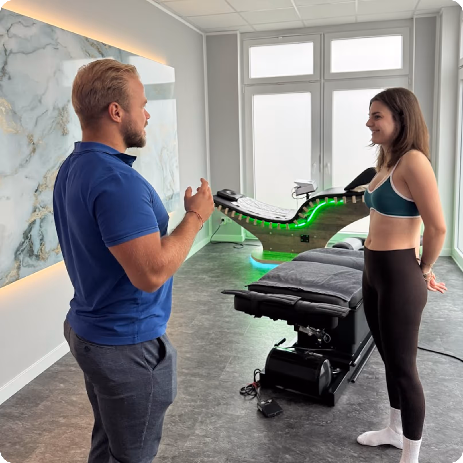 Chiropraktor im blauen Polohemd spricht mit einer Frau in Sportbekleidung in einem modernen Behandlungsraum mit Therapie- und Massageliegen.