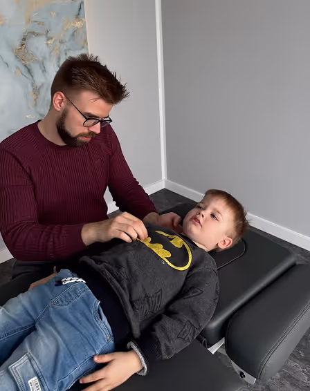 Ein Osteopath mit Brille und Bart untersucht einen Jungen, der auf einer medizinischen Liege liegt.
