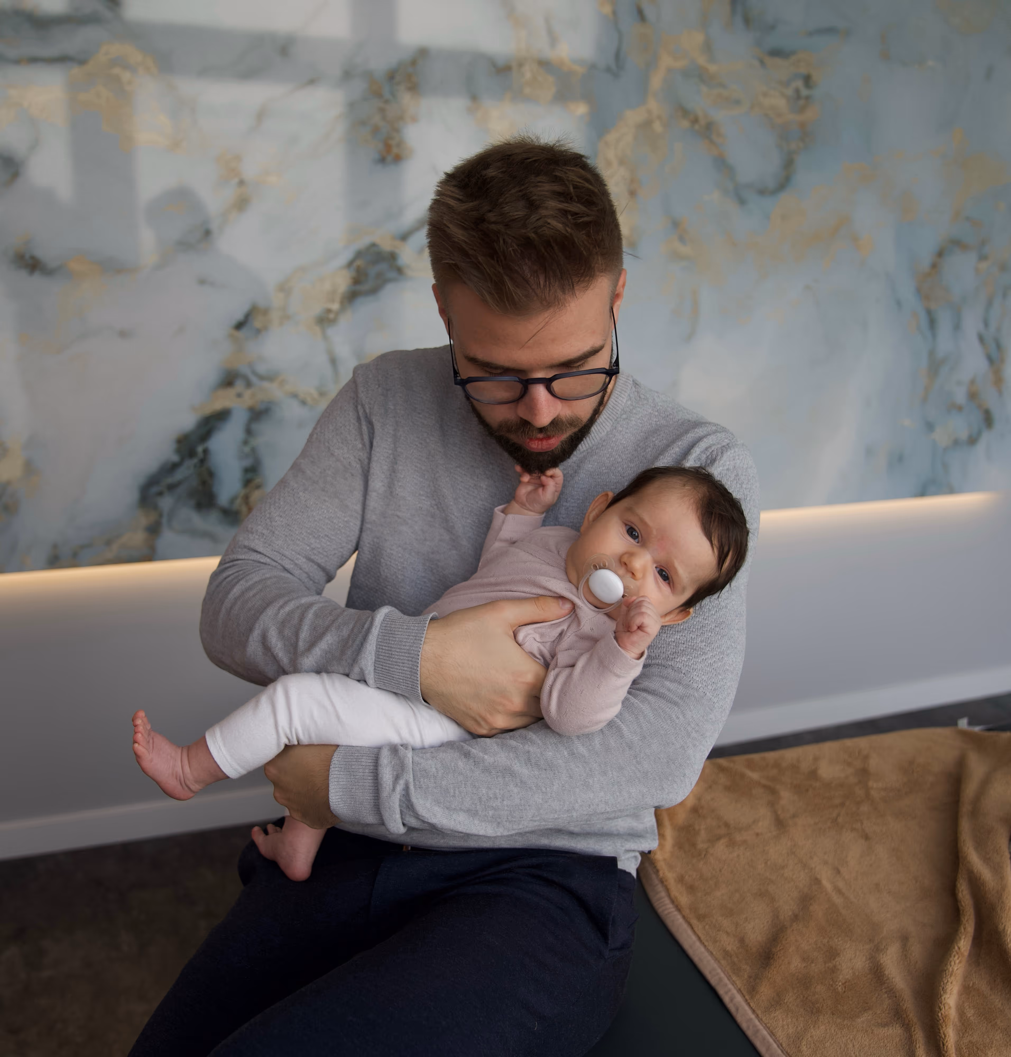 Ein Osteopath mit Brille und grauem Pullover hält ein Baby, das einen Schnuller hat und den Blick zur Kamera richtet.