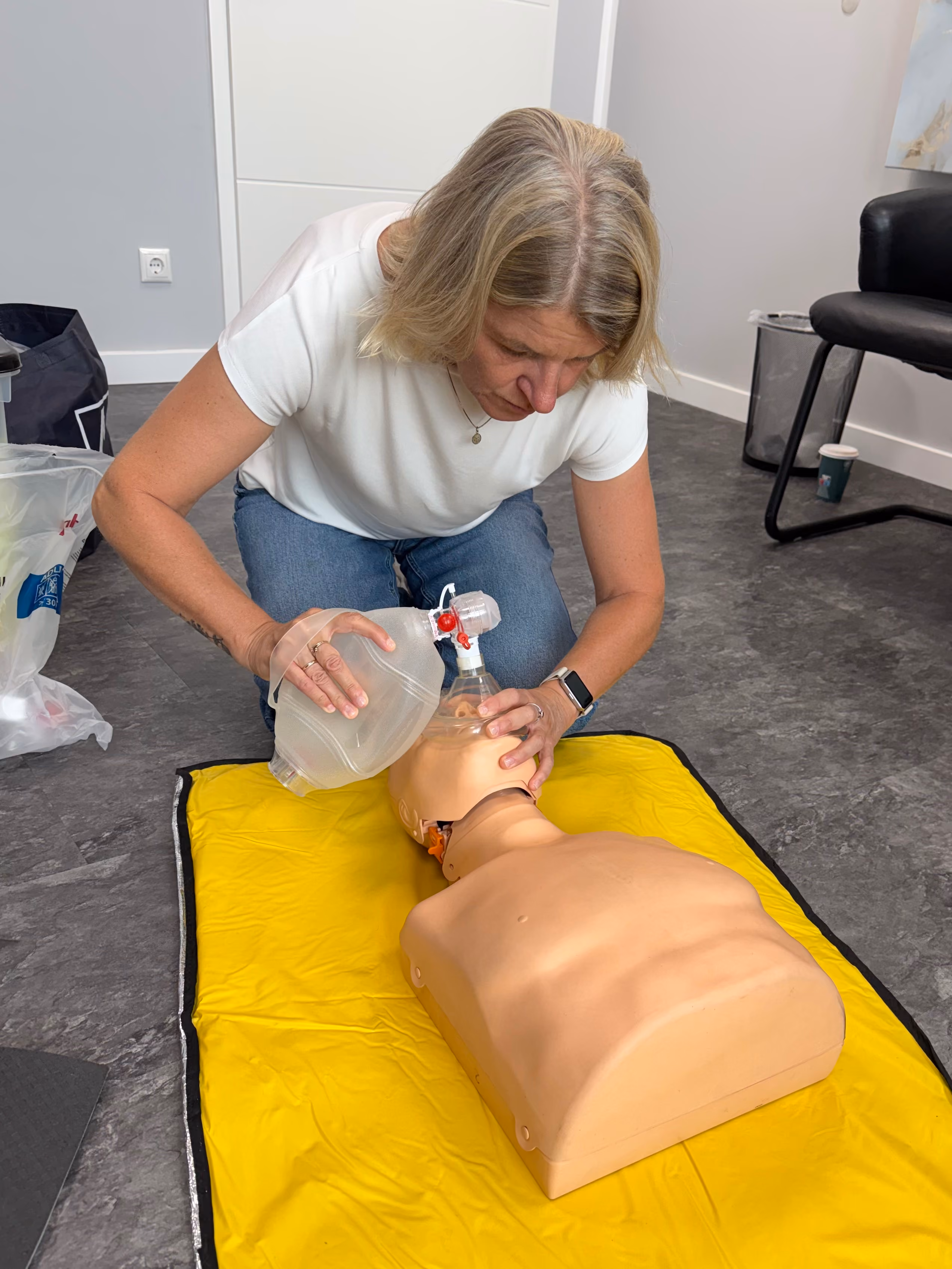 Frau übt Beatmung an einer CPR-Puppe auf gelber Matte in einem Raum mit Stuhl und Mülleimer.