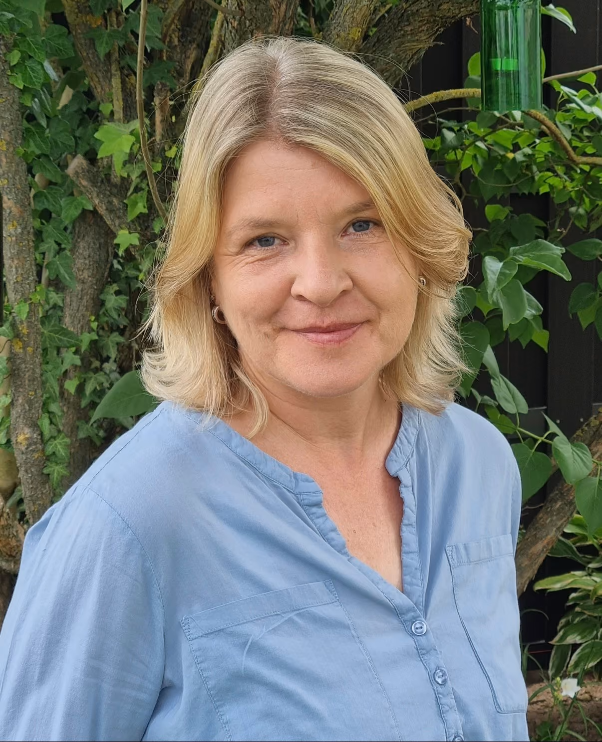 Frau mit blonden Haaren und blauem Hemd vor grünem Laub und Baumstämmen.