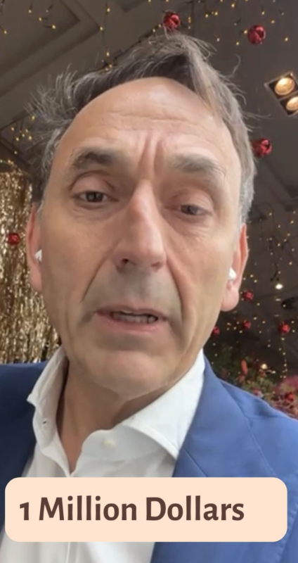 Close-up of a middle-aged man in a blue suit jacket and white shirt speaking, with festive decorations and red ornaments hanging in the background, and text overlay reading '1 Million Dollars'.