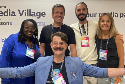 Five smiling people posing at a media village event, standing against a white background with visible name tags and lanyards.