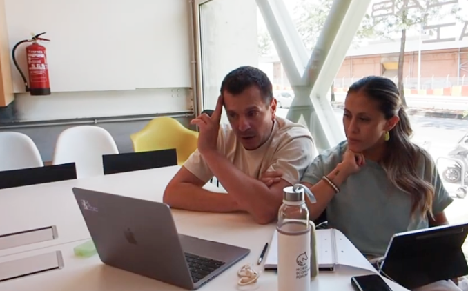 Two people sitting at a table with a laptop, tablet, notebook, and water bottle, engaged in discussion in a bright office.