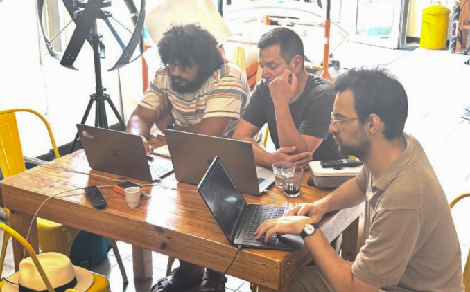 Three men sitting at a wooden table, working on laptops in a bright, casual office setting.