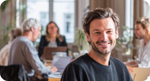 Smiling man in black sweater sitting in a bright office with colleagues working in the background.