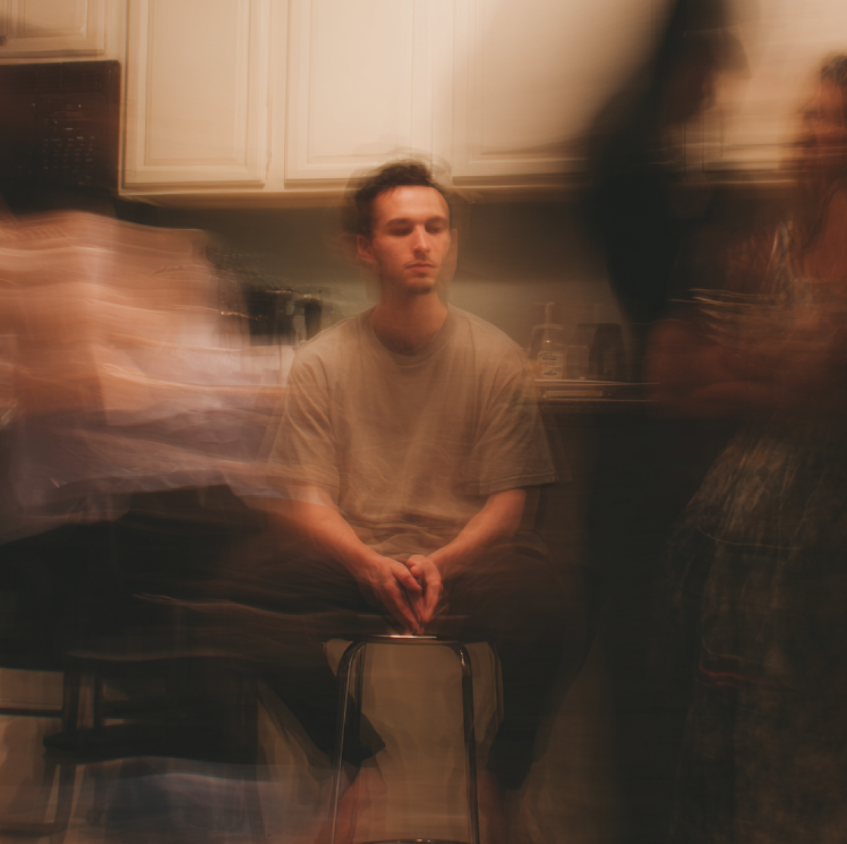 Young man sitting on a stool in a kitchen with blurred figures moving around him.