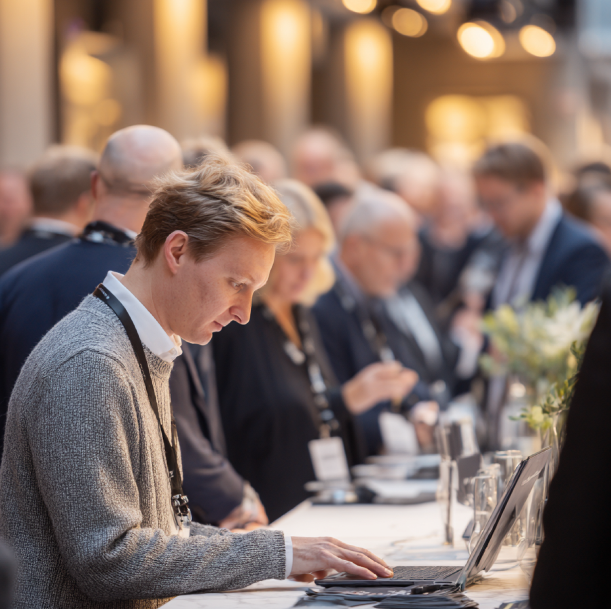 Young man in a gray sweater working on a laptop at a busy event registration table.