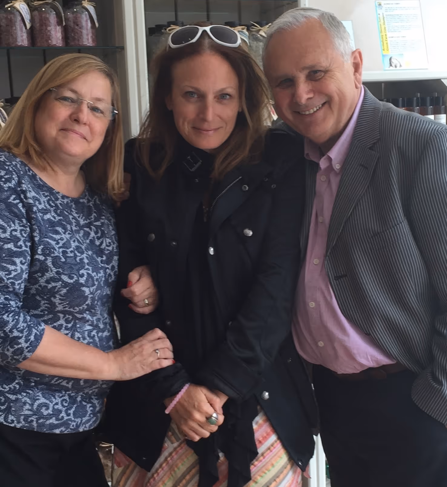 Three people standing close together smiling, with the woman in the center wearing sunglasses on her head and a black jacket.
