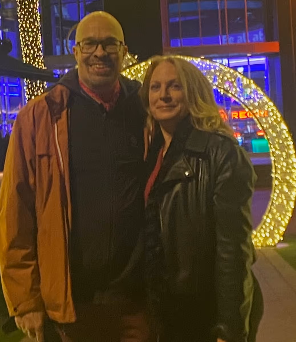 Smiling man in glasses and orange jacket standing next to a woman in a black leather jacket with illuminated decorative lights in the background at night.
