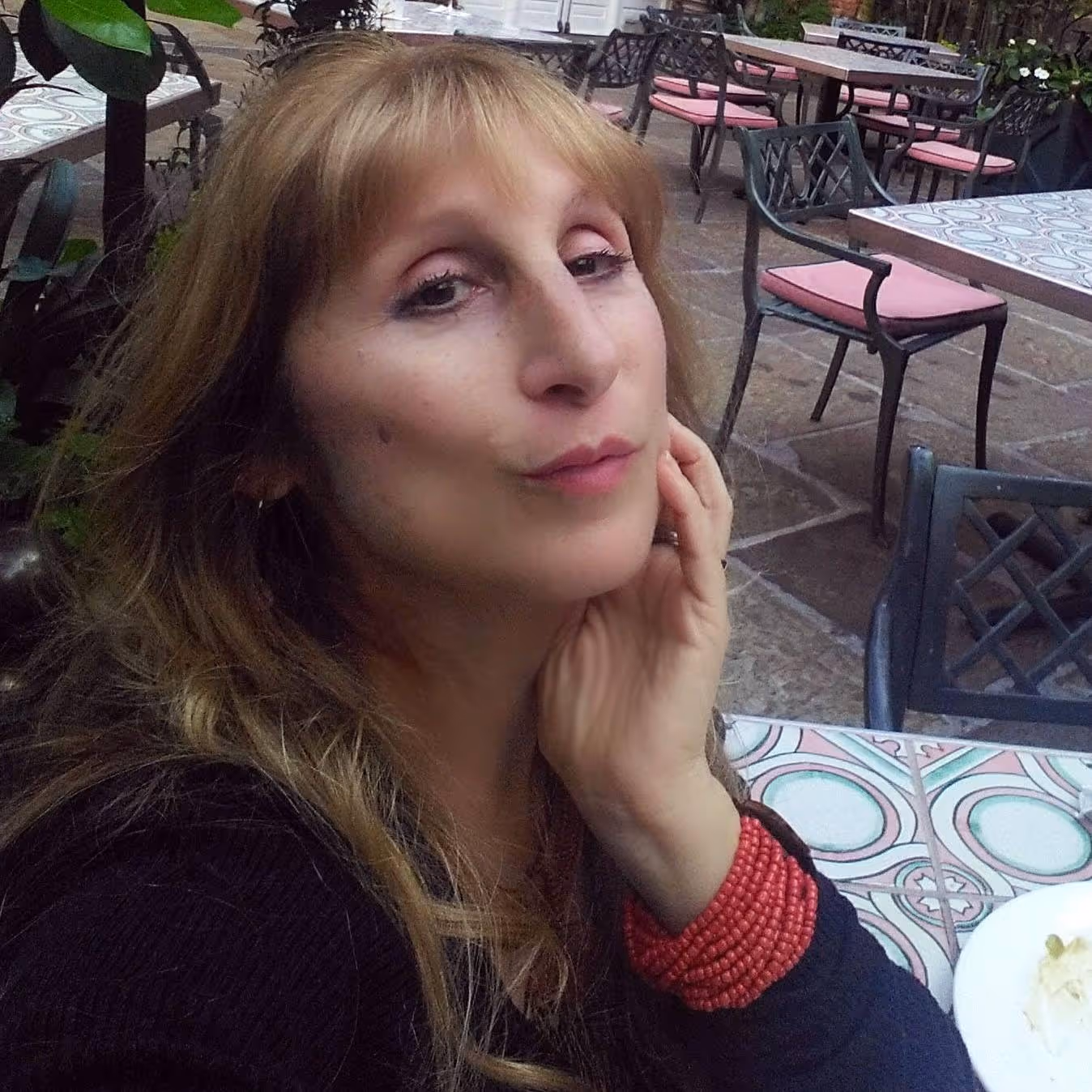 Woman with long blonde hair resting her hand on her cheek, sitting at a tiled outdoor café table with empty chairs in the background.