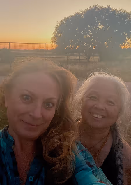 Two women smiling outdoors at sunset with a large tree and fence in the background.