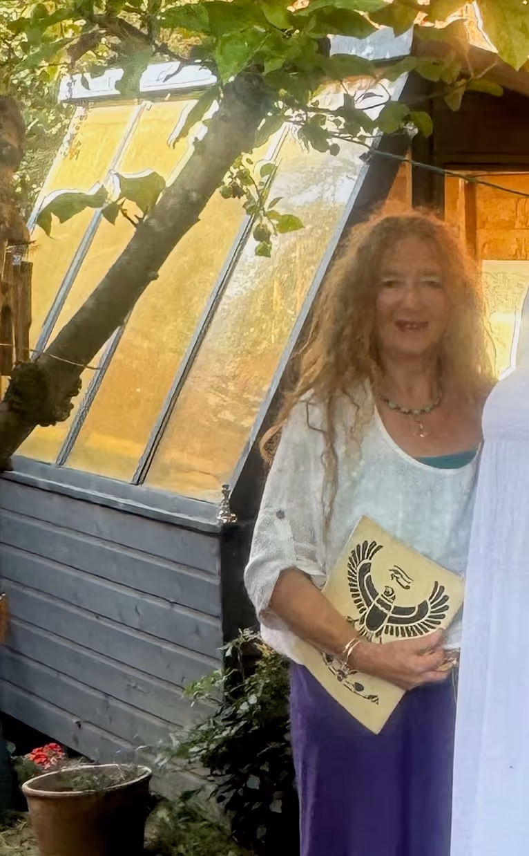 Woman with long curly hair standing outdoors near plants and a greenhouse, holding a folder with a black scarab beetle design.