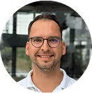 Mann mit Brille und weißem Hemd lächelt vor einem modernen Gebäude mit Glasfenstern.