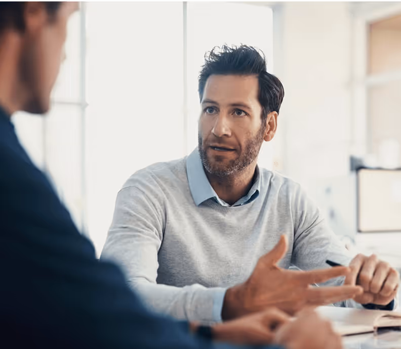 Zwei Männer im Gespräch, einer im Vordergrund mit Bart und grauem Pullover, gestikuliert in einem hellen Büro.