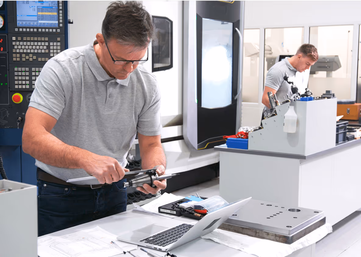 Mann nutzt Messschieber in moderner Werkstatt; Kollege arbeitet an Maschine im Hintergrund.