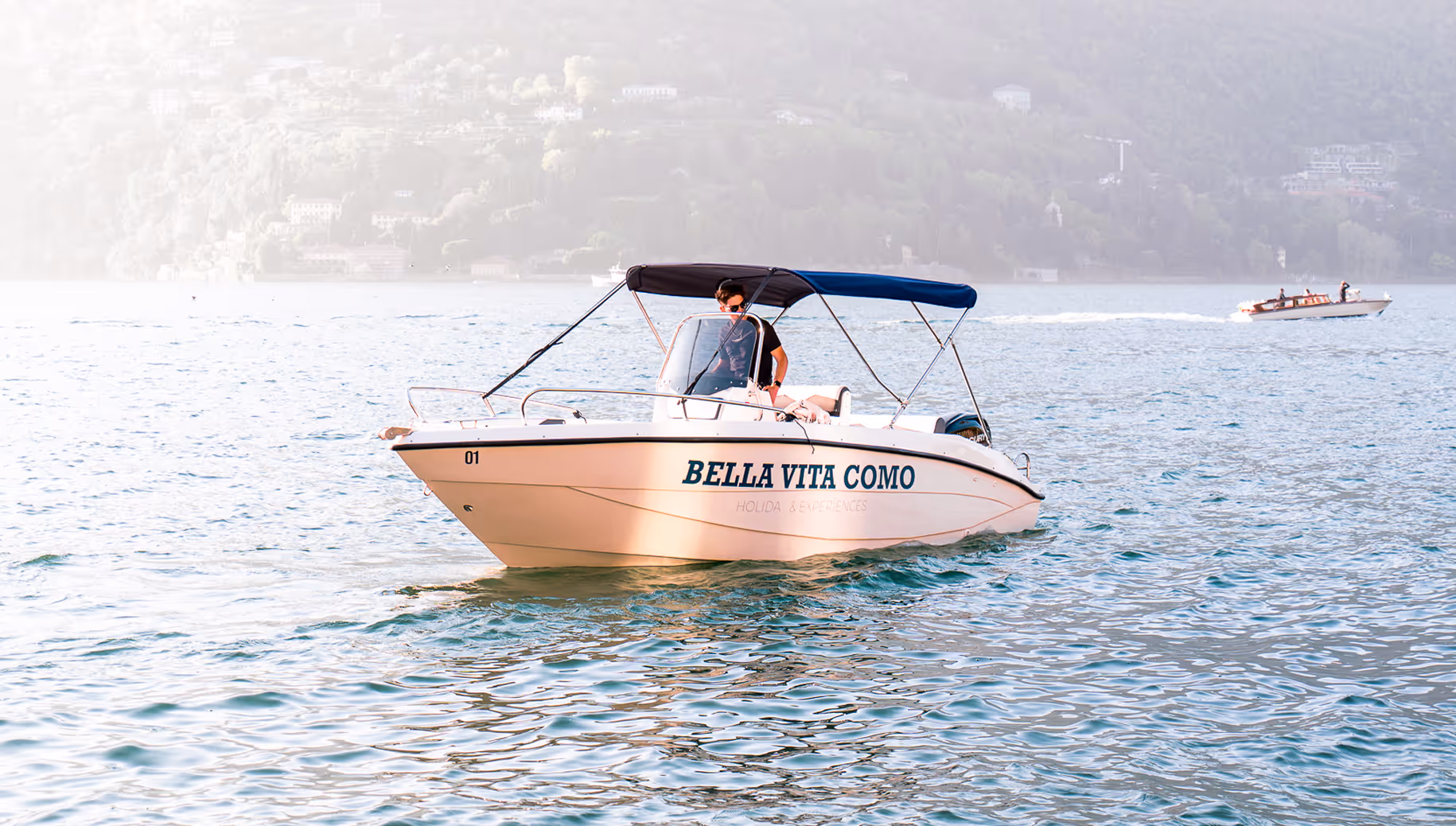 Маленькая белая моторная лодка под названием Bella Vita Como, на которой мужчина в солнцезащитных очках едет по спокойной воде на фоне туманного склона холма.
