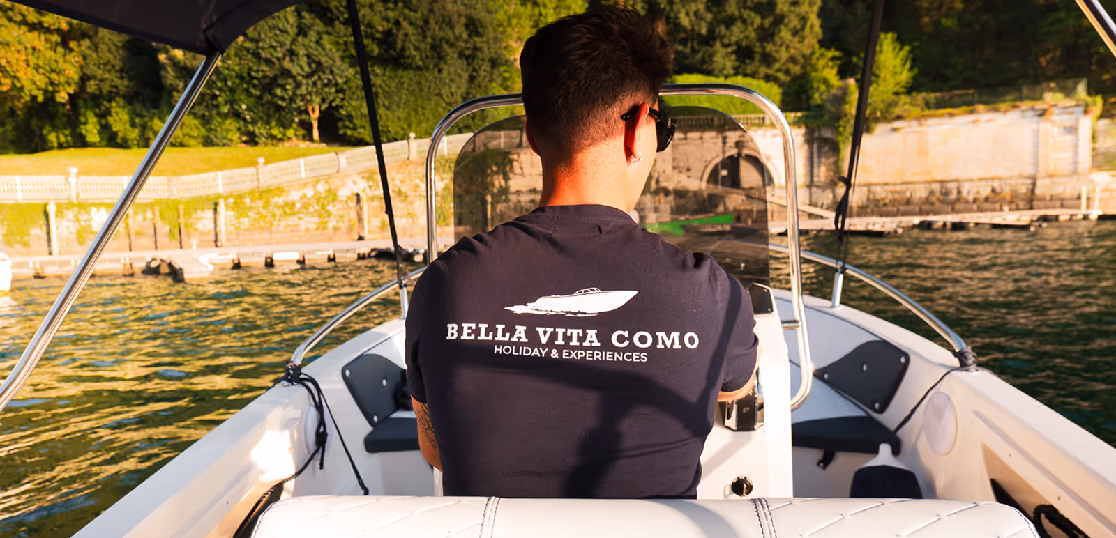 Человек с короткими волосами в солнцезащитных очках и футболке Bella Vita Como Holiday & Experiences управляет небольшой моторной лодкой по озеру рядом с каменным причалом и зелеными деревьями.