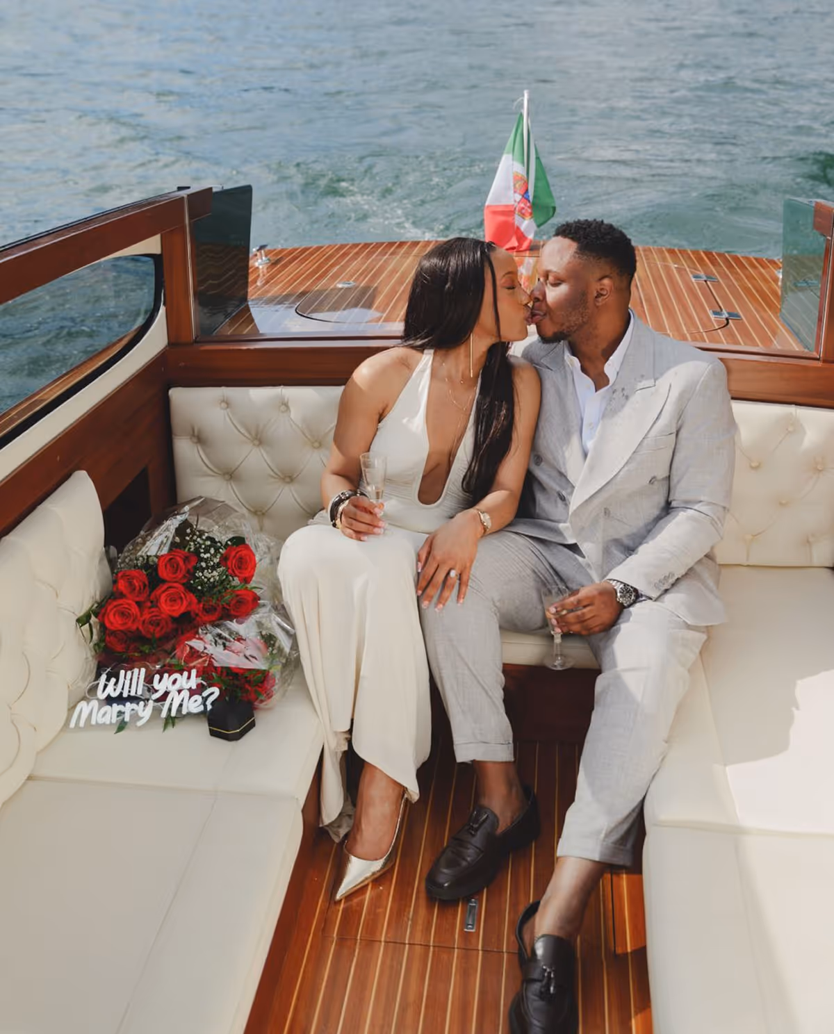 Couple sitting on a boat kissing, with a bouquet of red roses, a 'Will you marry me?' sign, and a ring box beside them.