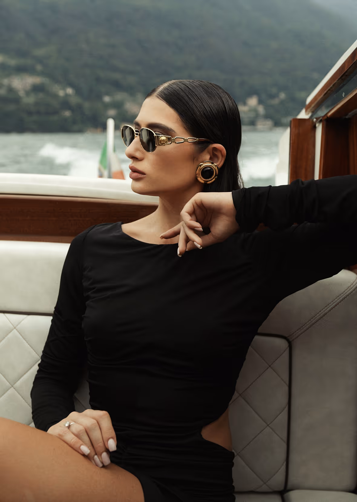 Woman wearing black dress and gold earrings, seated on a boat with water and mountains in the background.