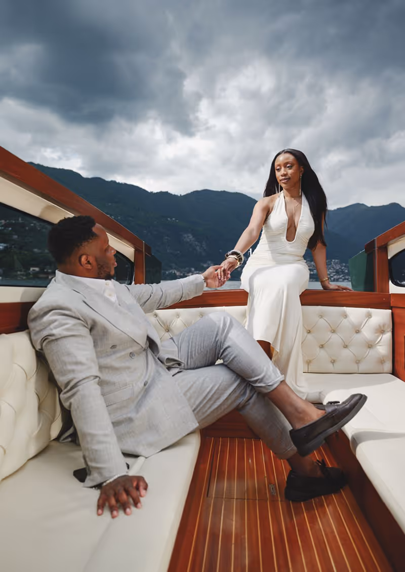 A couple dressed elegantly, sitting and holding hands on a boat with mountains and cloudy sky in the background.