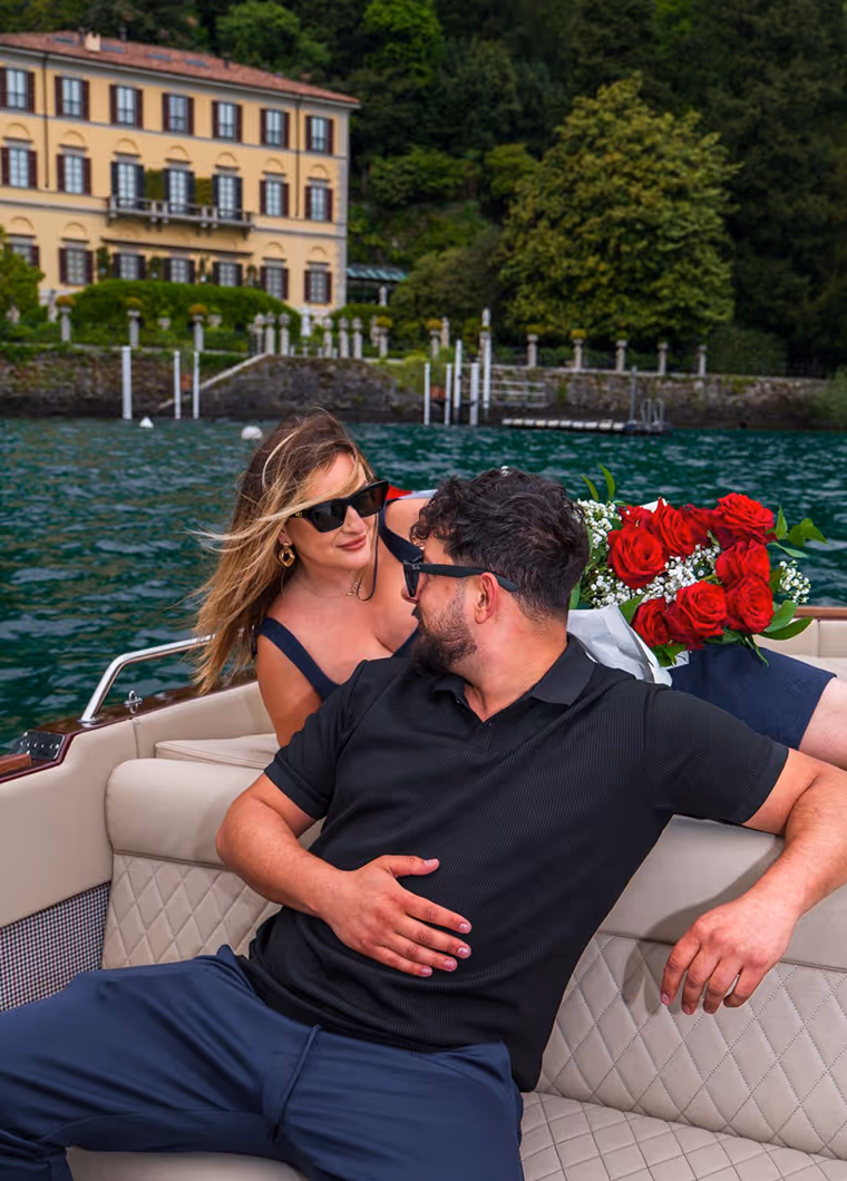 Couple wearing sunglasses sitting close together on a boat with red roses and a large house and green trees in the background on a lake.