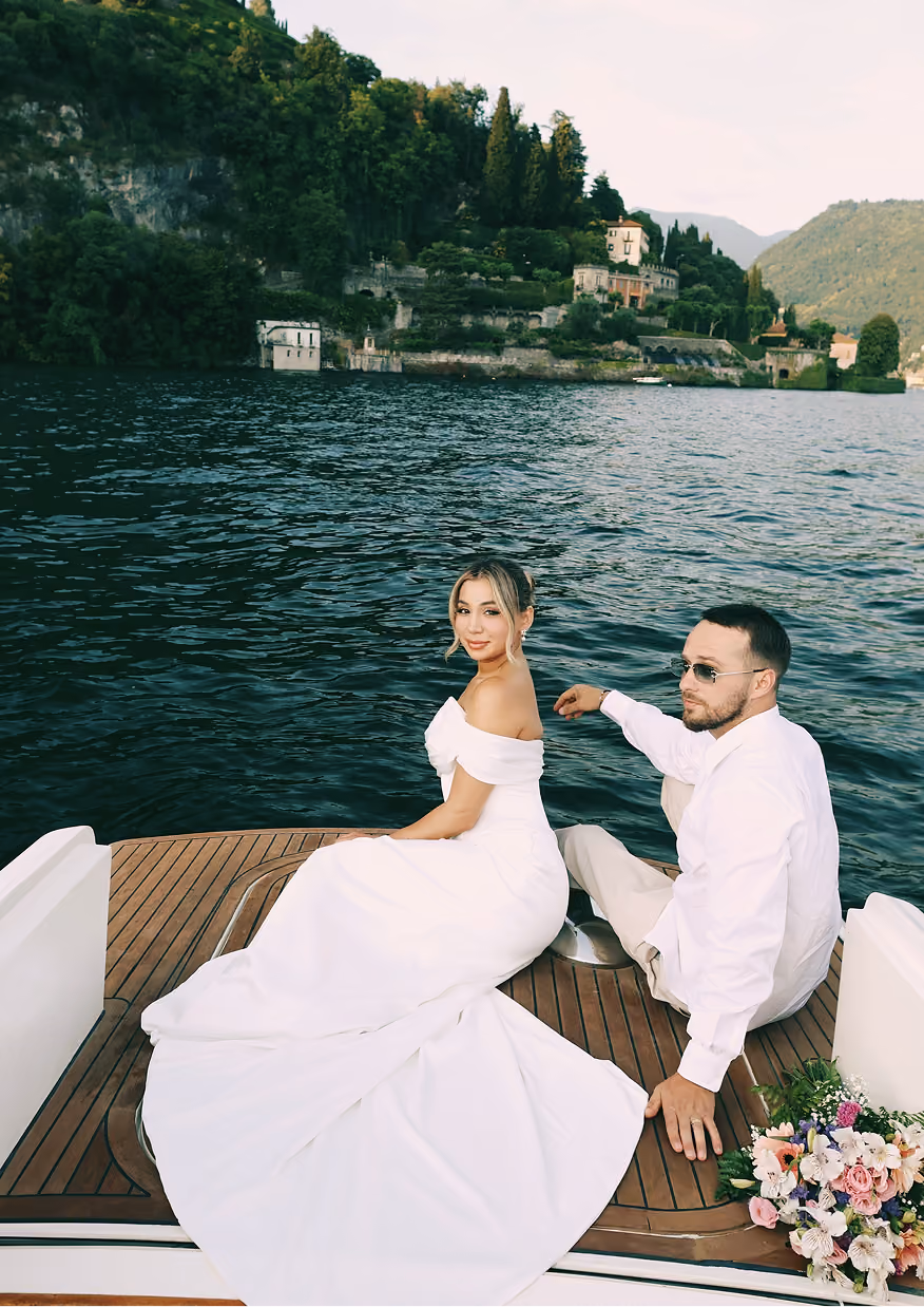 Woman in a glittery beige dress and gold high heels sitting on a boat with a scenic lakeside building and greenery in the background.
