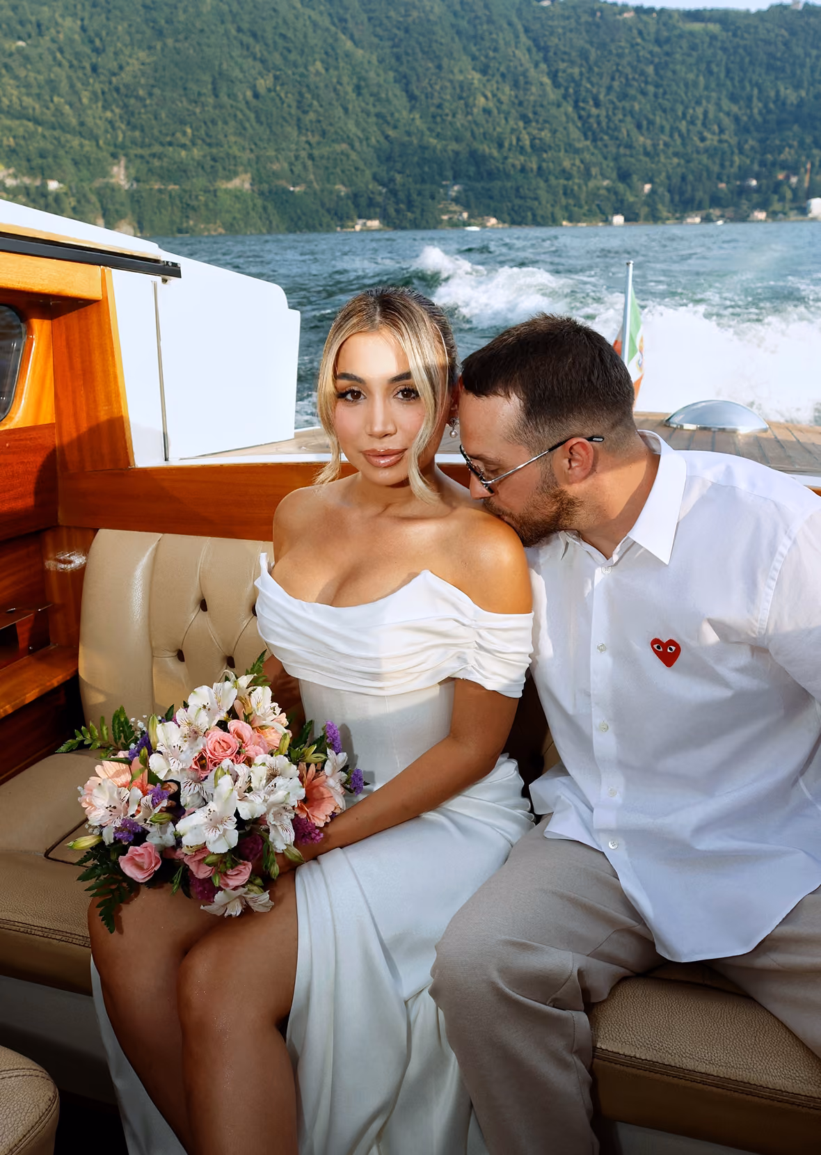 Woman in a white off-shoulder dress with pearl earrings posing on a boat with lake and green hills in the background.