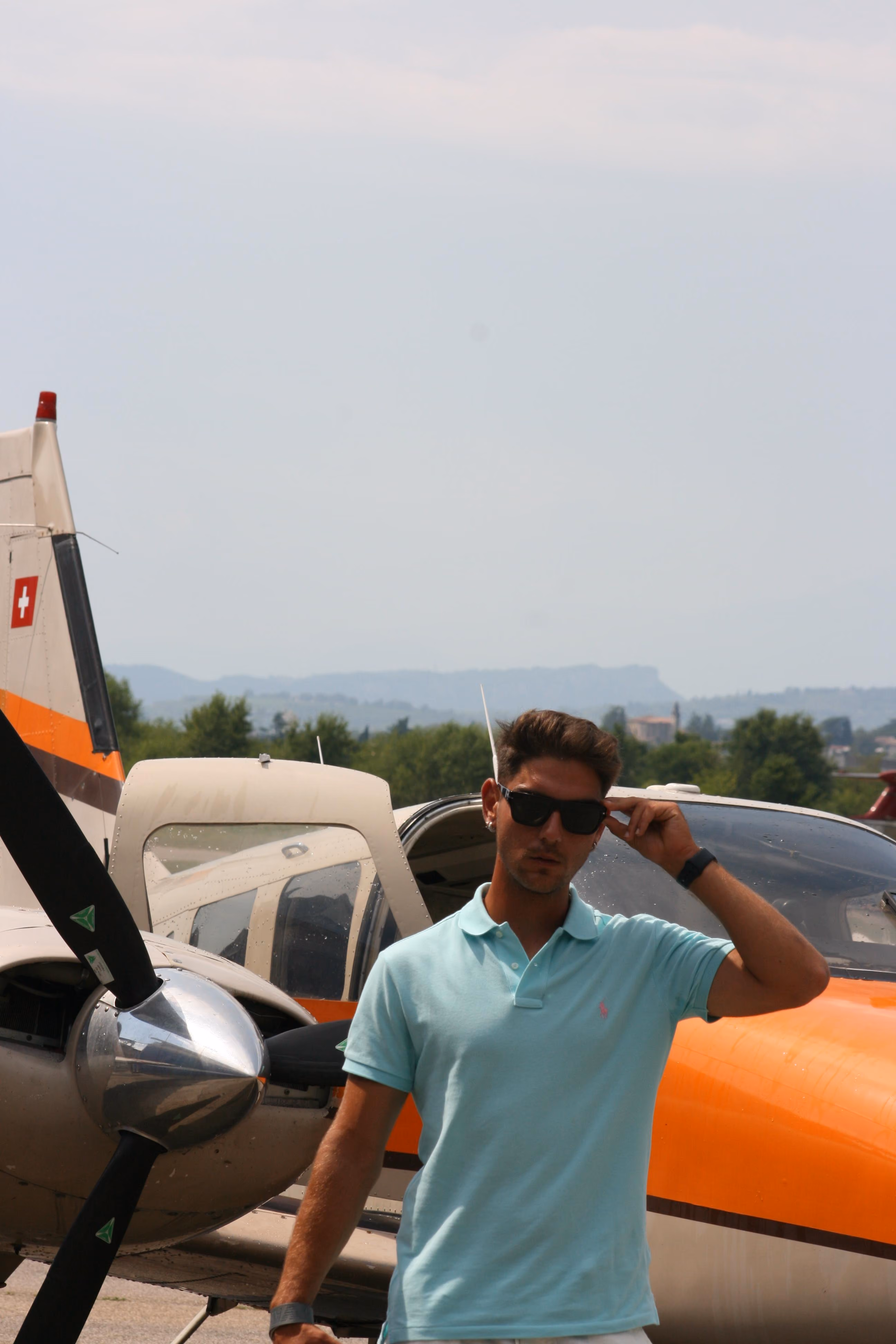Man wearing sunglasses and a light blue polo stands near a small orange and white airplane.