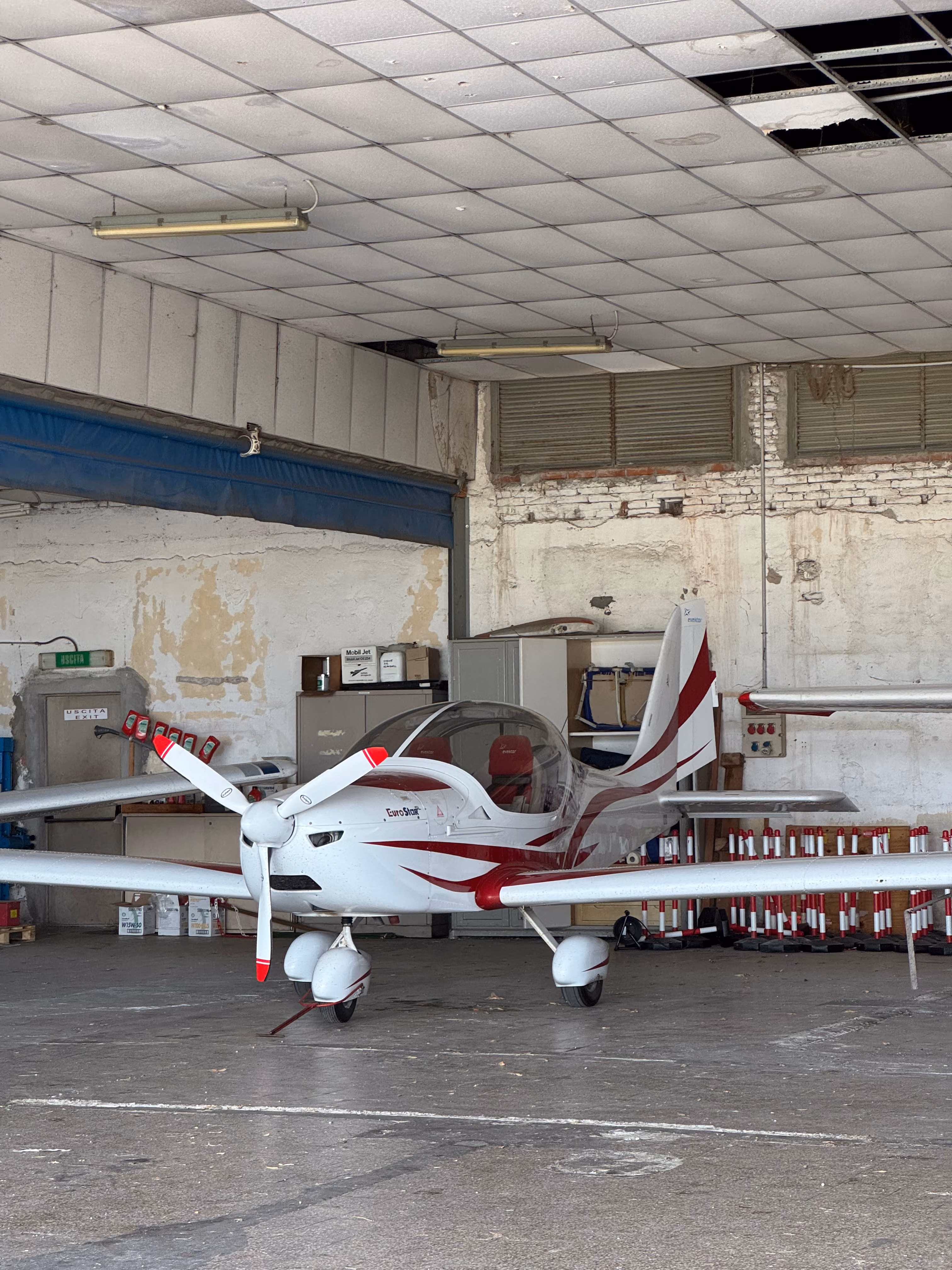 Small white and red EuroStar light aircraft parked inside a hangar with a damaged ceiling.