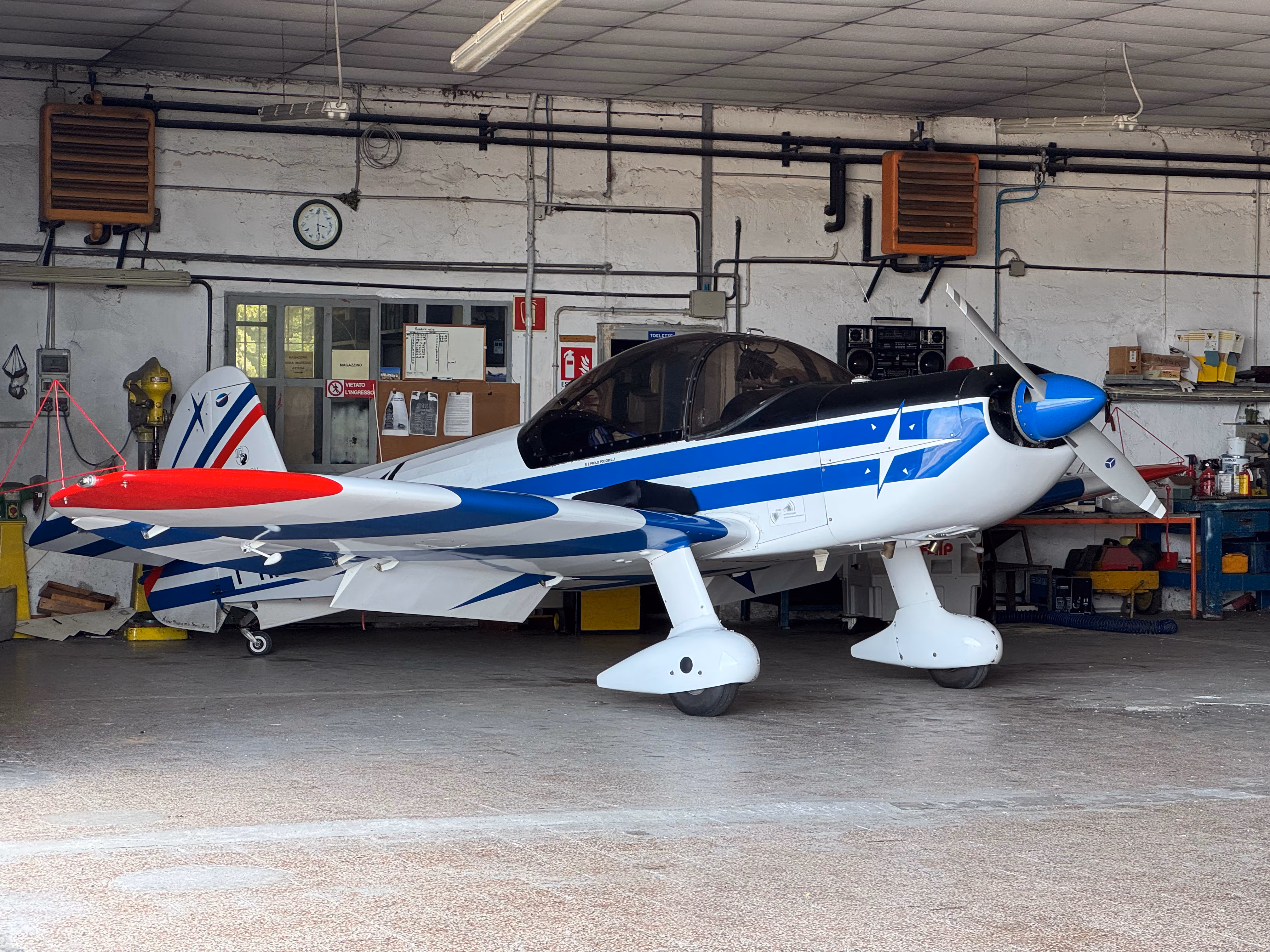 Small white airplane with blue stripes and red wingtip inside a hangar with tools and equipment.