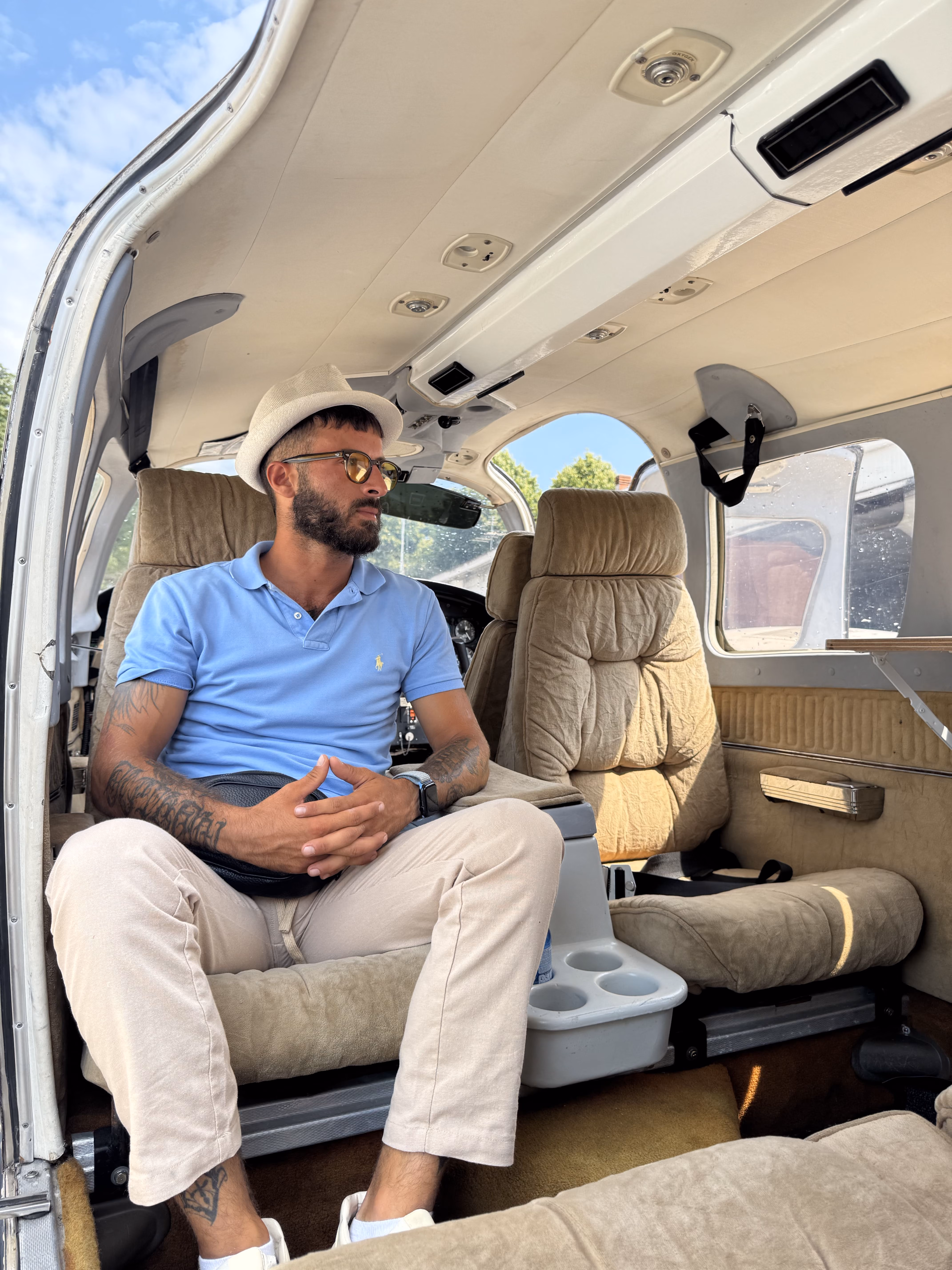 Man in a blue polo, beige pants, white hat, and sunglasses sitting inside a small airplane cabin.