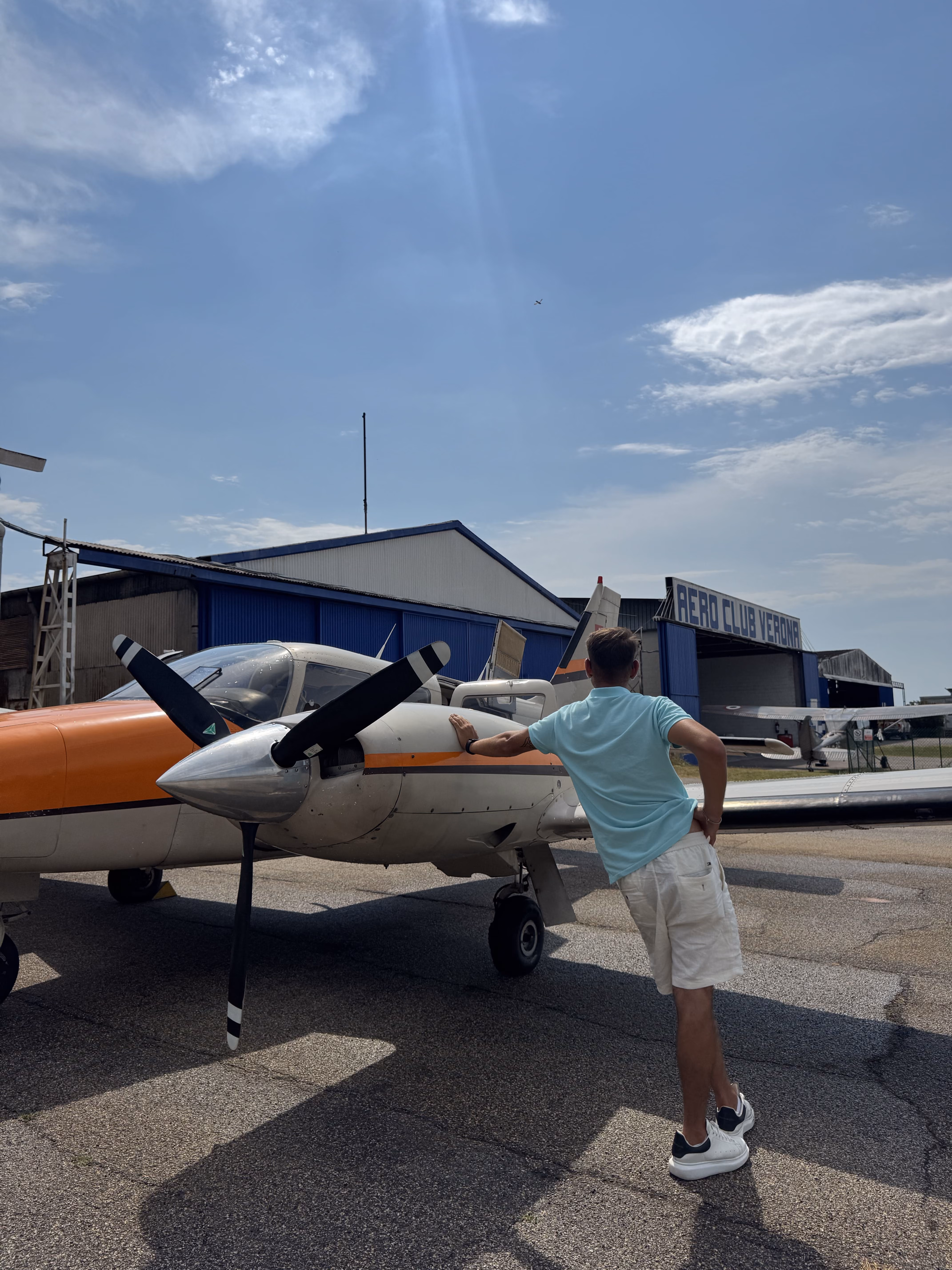 Man in blue shirt leaning on a small white and orange propeller plane outside a hangar.