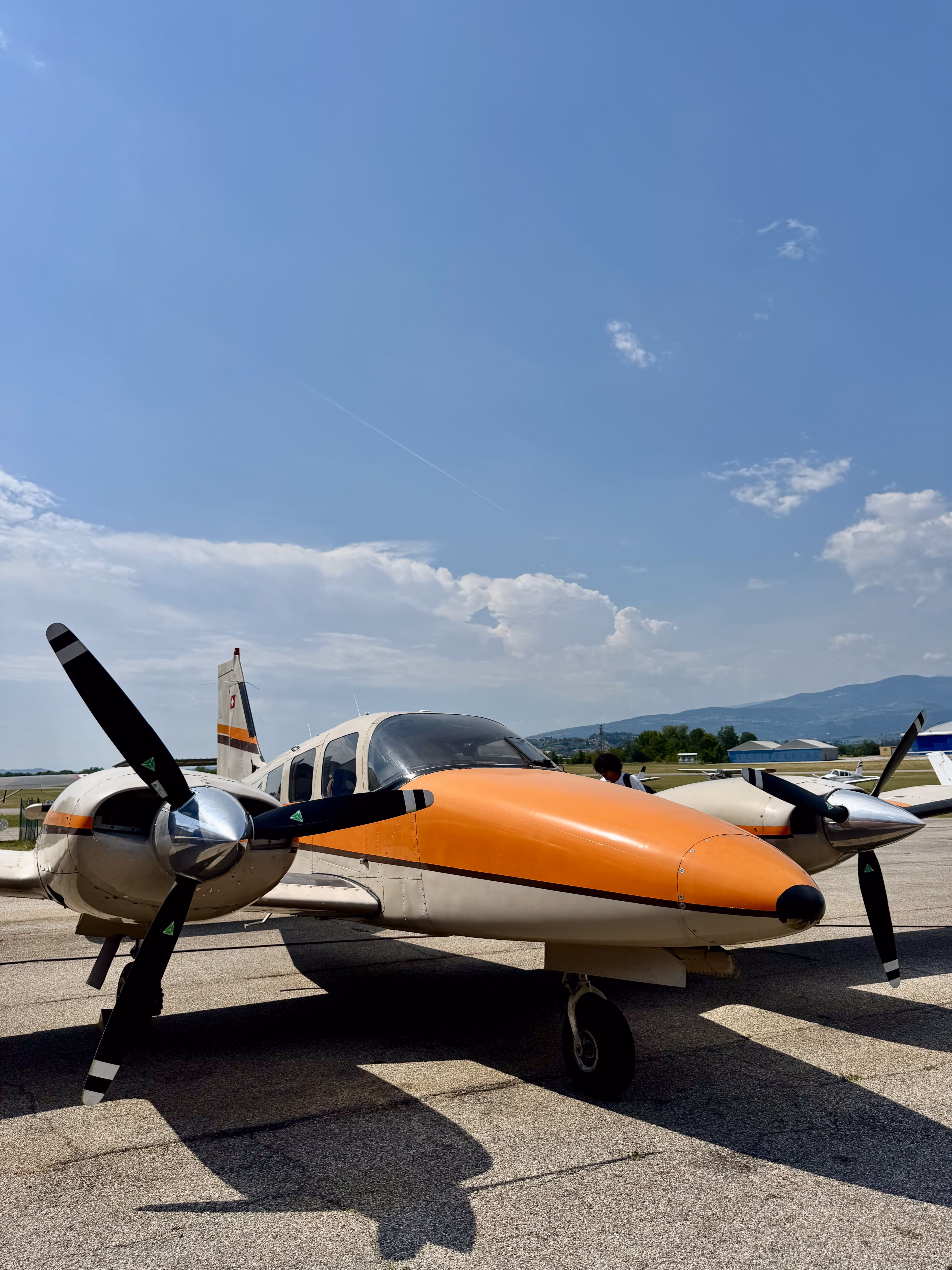 Small twin-engine propeller airplane with orange and white body parked on runway under a clear sky.