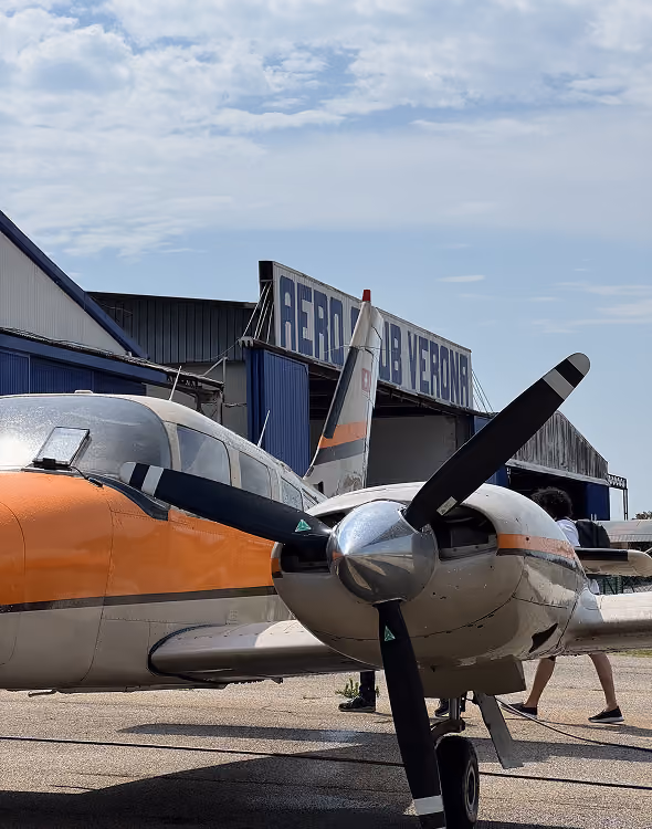 Small vintage airplane with orange and white paint parked outside hangar reading Aeroclub Verona.