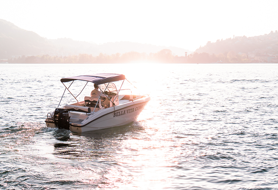 Small white motorboat with canopy on sparkling lake water at sunset with distant hills and trees.