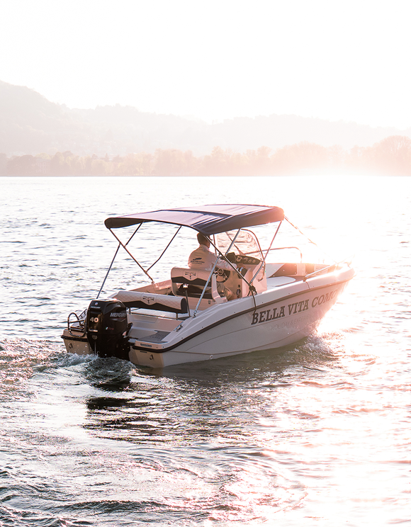 White motorboat named Bella Vita Comodo on water with a person wearing a cap driving at sunset.