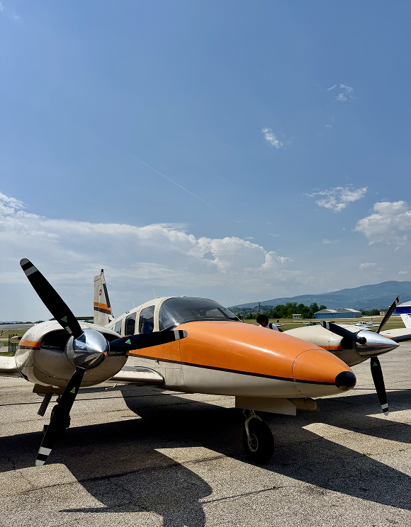 Small twin-engine propeller airplane with orange nose parked on runway on clear day.