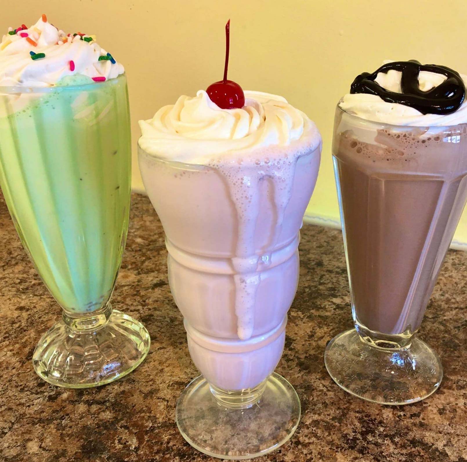 Three colorful milkshakes in tall glasses are displayed on a counter, each topped with whipped cream.
