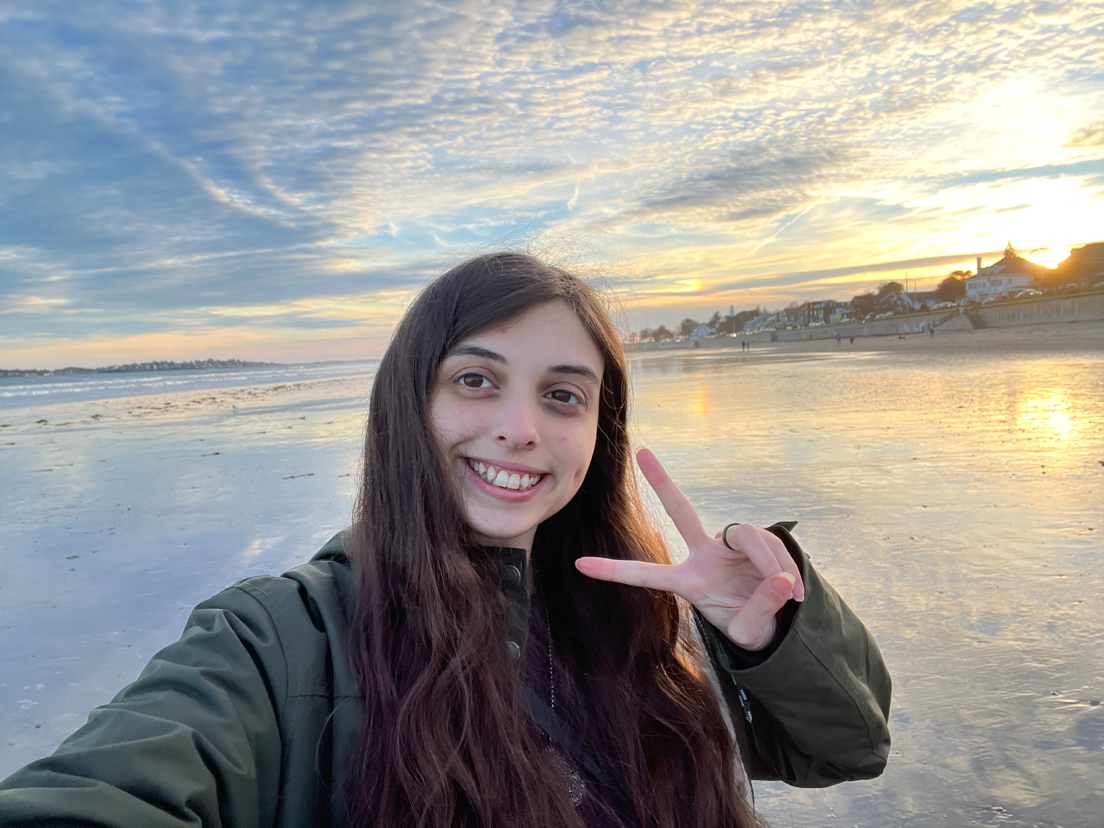 A photo of Rhi - a young woman with long dark hair smiling and making a peace sign on a sandy beach at sunset.
