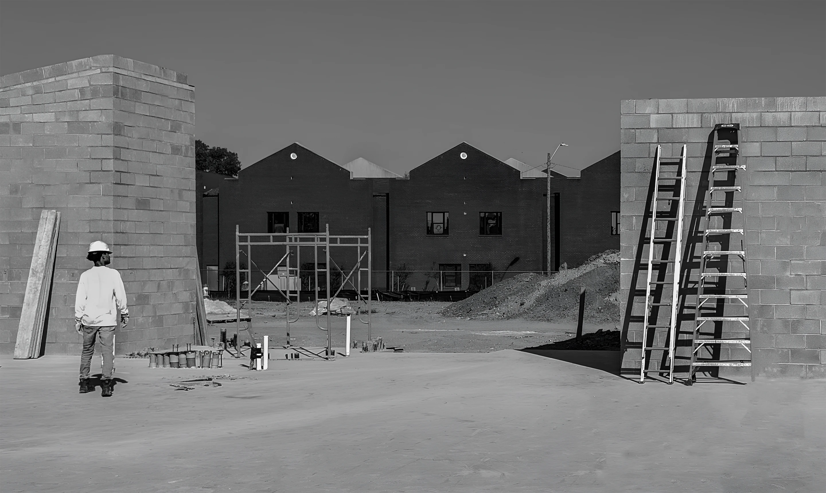 Construction site with a worker wearing a helmet, concrete walls, ladders leaning against a wall, and scaffolding in front of brick buildings.