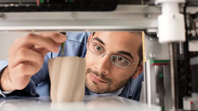 Man wearing glasses closely inspecting a 3D printed mug inside a 3D printer.