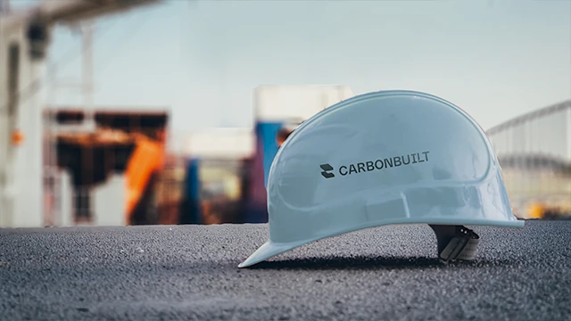 White construction hard hat with Carbonbuilt logo resting on a textured surface with a blurred construction site background.