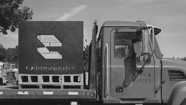 Flatbed truck carrying a large metal block with the word CARBONBUILT and a stylized C logo cut out.