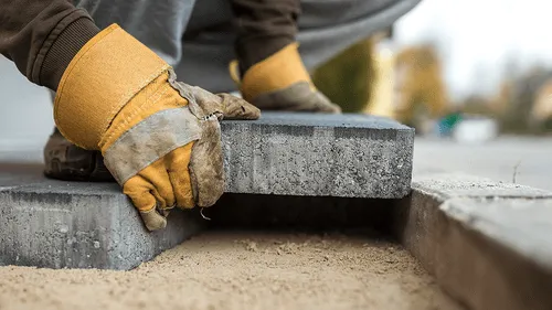 Person wearing gloves placing a gray paving stone onto a sand base during outdoor pavement installation.