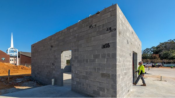 Construction worker walking beside an unfinished concrete block building on a sunny day.