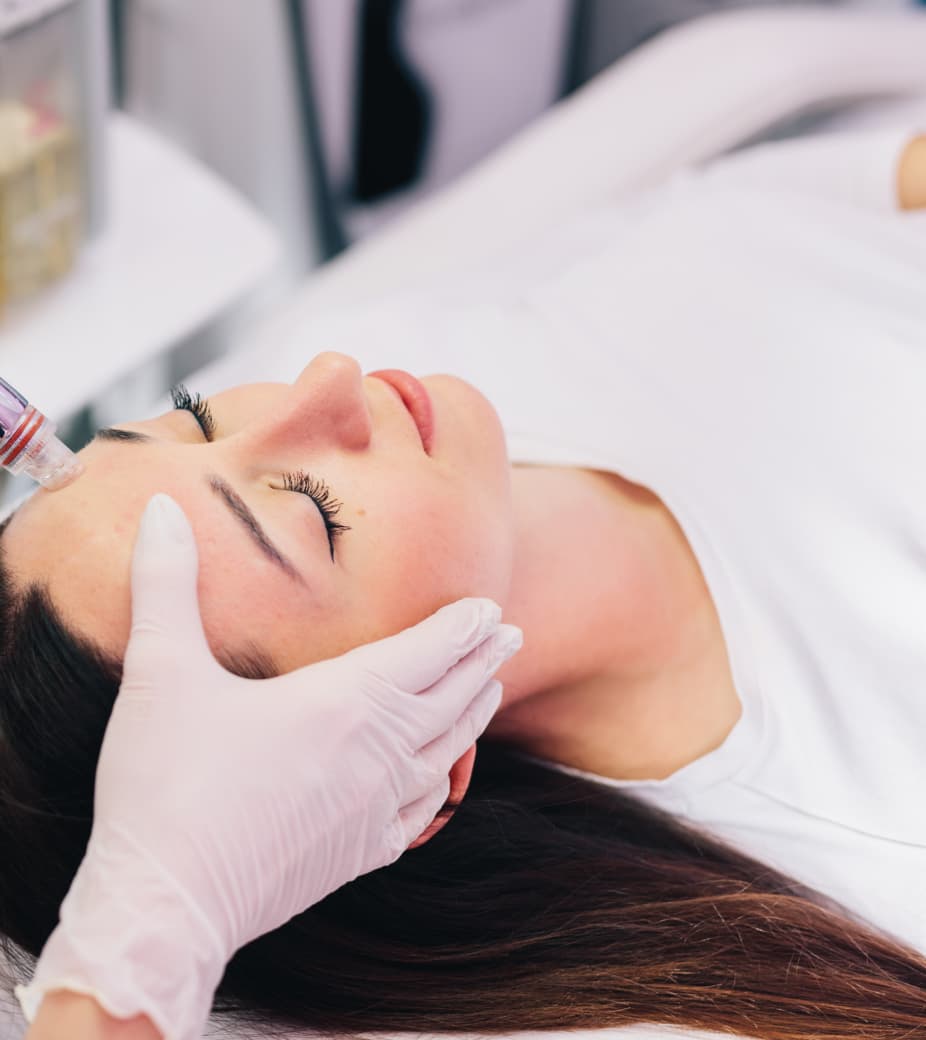 Woman receiving a facial treatment with a microneedling device applied to her forehead by a gloved hand.