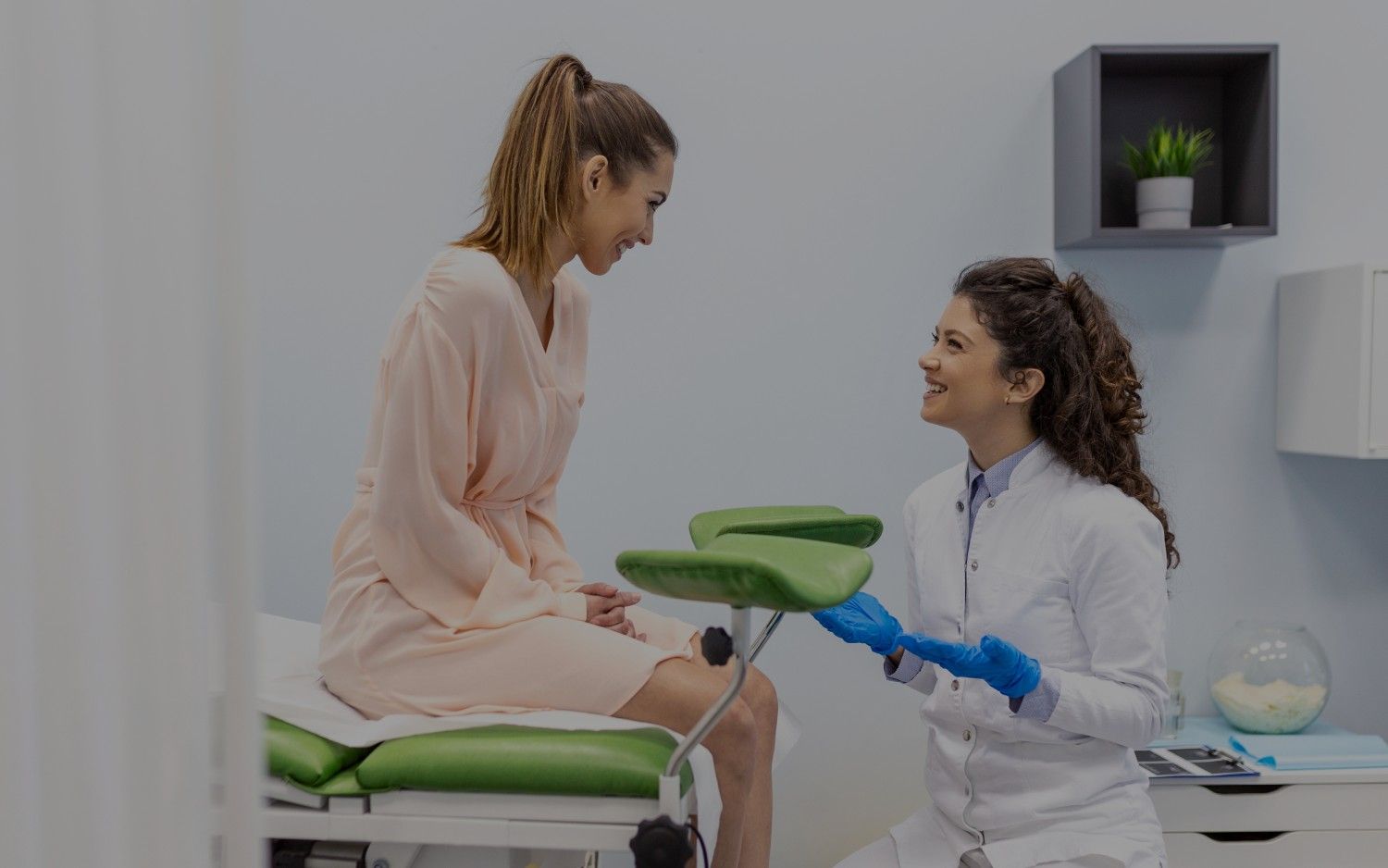 Female doctor wearing blue gloves smiling and talking to a female patient seated on a green examination chair in a medical office.