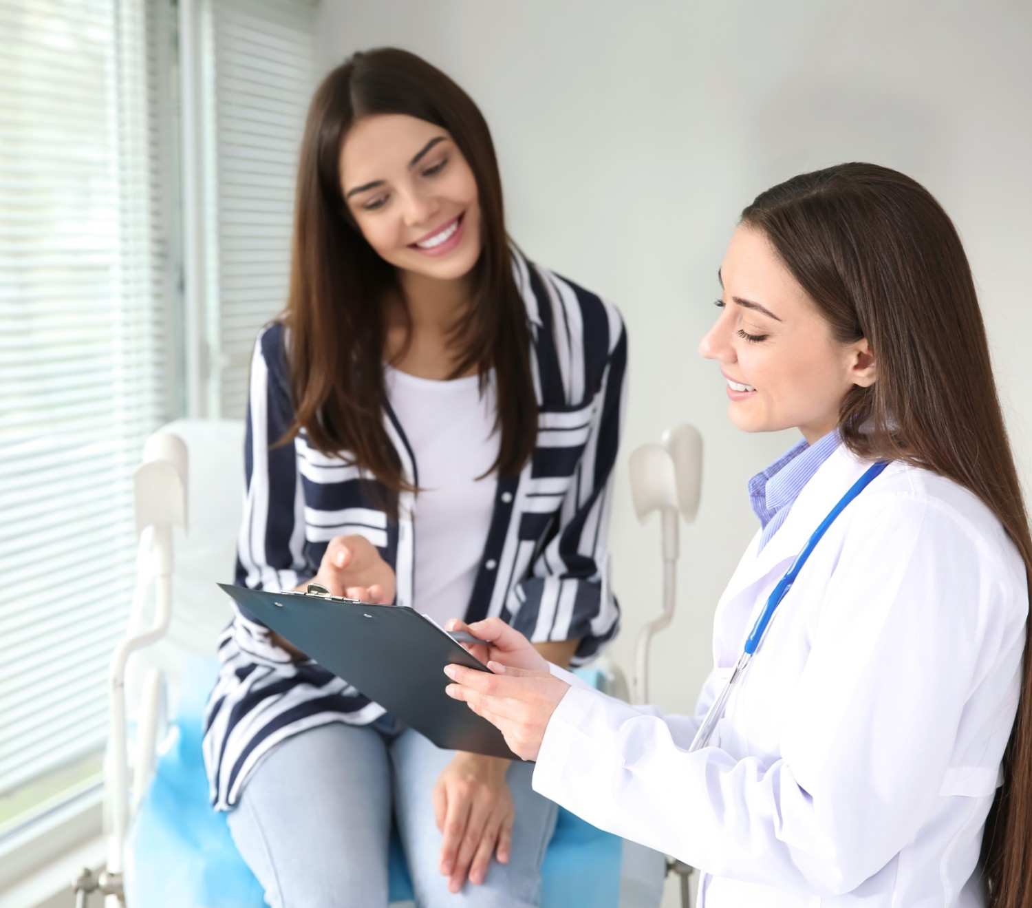 Female doctor showing a clipboard to a smiling patient sitting on a medical chair in a clinic.